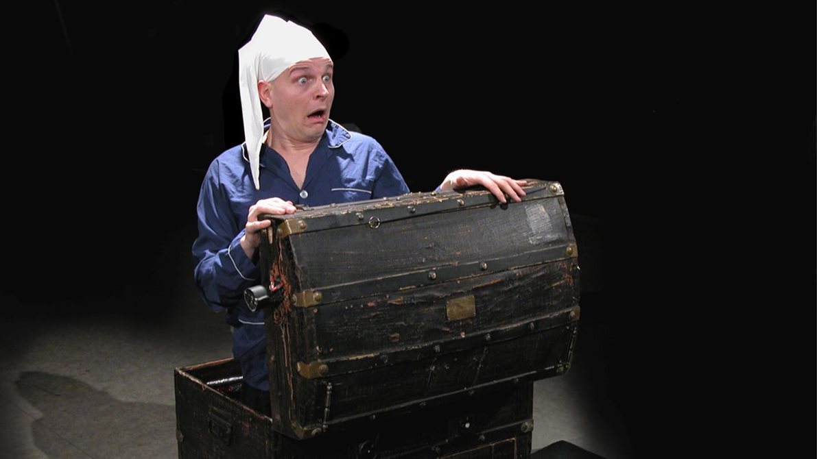 En mann i en kiste titter opp med en forskrekket mine i full pysj og sovelue
