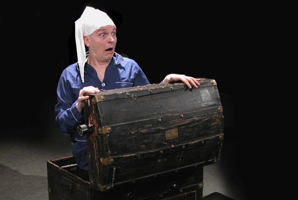 En mann i en kiste titter opp med en forskrekket mine i full pysj og sovelue