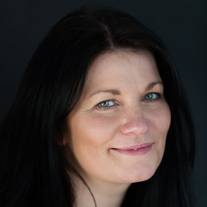 Merete Bø ser i kameraet med hodet litt på skjeve. Hun har mørkt, utslått hår og smiler med lukket munn
