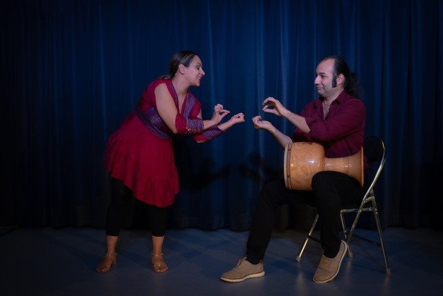 En mann og en kvinne er på en scene. Mannen sitter på en stol med en tromme på fanget, mens kvinnen står ved siden av vendt halvveis mot han. De begge holder tommel og pekefinger sammen på begge hendene.